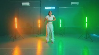 Renkli ışıkları ve arka planda neon ışığı olan bir stüdyoda enerjik bir şekilde dans eden bir kadın..