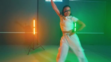 Renkli ışıklı ve neon ışıklı bir stüdyoda dans eden biri. Stüdyo, aydınlatma armatürleri ve düz zemini olan modern bir tasarıma sahip..