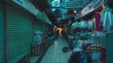 Bir tarafında kapalı dükkanlar ve diğer tarafında plastik eşyalar satan birkaç açık tezgah olan loş bir pazar yeri. Ara sokağa bir motosiklet park etmiş..