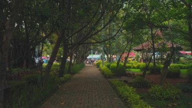 Bereketli yeşillikler ve ağaçlarla çevrili sakin bir park yolu, sağda bir çardak ve arka planda insanlar var..