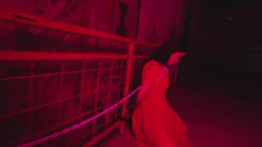 Uzun saçlı bir insan loş, kırmızı renkli bir ortamda duruyor. Omuzlarında bir zincir var ve aşağı bakıyor gibi görünüyorlar..