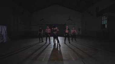 Dört kişilik bir grup, loş, geniş bir kapalı alanda dikiliyorlar. Koyu renk, gergin kıyafetler giymişler ve rol yapıyor ya da poz veriyor gibi görünüyorlar. Yerde uzun gölgeler var..