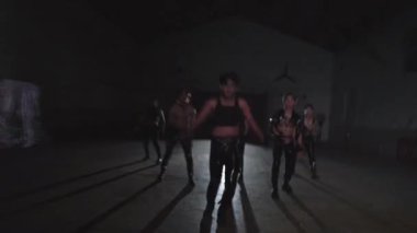 Bir grup dansçı loş bir alanda enerjik bir şekilde performans sergiliyorlar, koyu, parlak kıyafetler giyiyorlar..