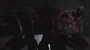 Bir grup genç adam karanlık, tehlikeli kıyafetler ve zincirler içinde loş bir endüstriyel ortamda dramatik bir dans gösterisi yapıyorlar..