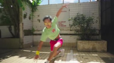 Yeşil tişörtlü ve kırmızı şortlu bir kişi arka planda bir ağaç ve bir duvarla açık alanda fayanslı bir zeminde çömeliyor..