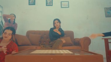 Oturma odasında kahverengi bir kanepede oturan ve önünde kahve masası olan iki kadın. Ön plandaki kadın bacak bacak üstüne atmış otururken, arka plandaki kadın kısmen görülüyor. Arkalarındaki duvarda çerçeveli resimler var..