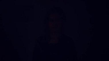 Ciddi bir ifade takınan, siyah giysili bir kadın, sade bir arka plana karşı içeride duruyor..