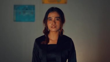 Ciddi bir ifade takınan, siyah giysili bir kadın, iki bulanık tabelalı düz bir arkaplanda duruyor..