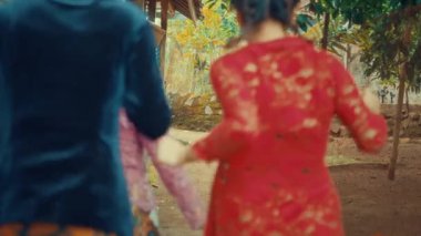 Kırsal alanda dar bir yolda yürüyen geleneksel kıyafetler giyen iki kadın. Bir kadın kırmızı dantelli bluz giyiyor ve diğeri koyu mavi bluz. Arkaplanda ağaçlar ve eski bir bina var..