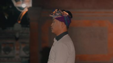 Geleneksel kıyafetler ve renkli bir başlık takmış loş bir ortamda yan yana duran bir adam..