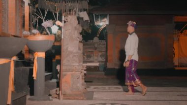 Geleneksel giysiler giymiş bir kişi bir tapınak ya da tapınağın önünde duruyor, muhtemelen kültürel ya da dini bir törene katılıyor. Dekoratif unsurları ve sunumları olan bir açık hava alanı gibi görünüyor..