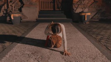 Geleneksel giyinmiş bir kişi bina girişinin önündeki çakıllı yolda eğilmiş, her iki tarafında saksı bitkileri olan biri..