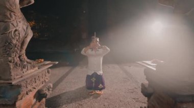 Diz çöküp kafasının üstünde tahta bir nesne tutan bir insan. Geleneksel bir ortamda, taş heykeller ve sisli bir atmosferde..