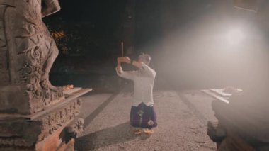 Geleneksel giysiler içinde bir ayin yapan ya da loş bir tapınak ortamında dans eden, her iki tarafında taş heykeller olan bir kişi..