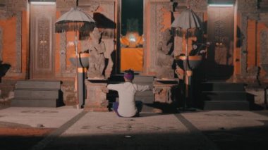 Geceleri Bali 'nin geleneksel bir tapınağının önünde kolları çapraz oturan sıcak ışıkla aydınlatılan bir insan. Tapınakta karmaşık oymalar ve heykeller var..