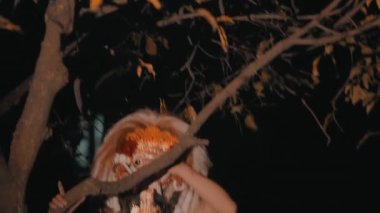 Geleneksel bir maske ve kostüm giyen bir insan, bir ağaç dalı tarafından kısmen gizlenmiş, karanlık bir açık hava ortamında.