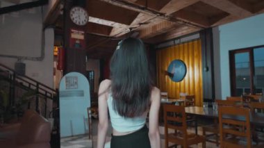 Uzun saçlı bir kadın bir kafede kameradan uzak duruyor. Beyaz bir bluz ve siyah bir etek giyiyor. Kafede ahşap mobilyalar ve kadını yansıtan büyük bir ayna var..