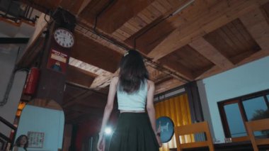 Uzun saçlı bir kadın endüstriyel tarzda bir odada duruyor. Tavanı ahşap ve boruları açıkta..