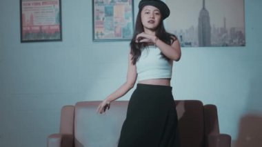 Bere takan ve beyaz bir bluz giyen genç bir kadın, arkasında çerçeveli posterlerle koltuğun önünde güvenle poz veriyor..