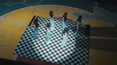 Yukarıdan bakıldığında, kareli bir zeminde bir sıra halinde duran altı kadından oluşan bir grup. Zeminde çatlaklar, sarı ve turuncu renklerin karışımı olan büyük dairesel bir dizayn var..