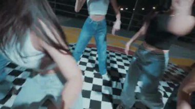 Bir grup genç kadın gece kareli zeminde enerjik bir şekilde dans ediyor. Günlük kıyafetler giyiyorlar. Ekin bluzları ve kotları da dahil. Arka plan karanlık ve bazı ışıklar görülebilir..