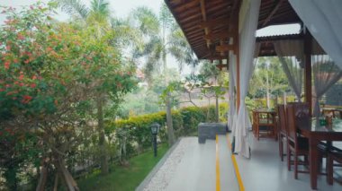 Çiçek açan bir bahçenin, bir patikanın ve ahşap bir avlunun manzarası. Veranda, yemyeşil ve palmiye ağaçlarıyla çevrili ahşap mobilyalarla dolu sakin bir atmosfer yaratan bir yemek alanına sahip..