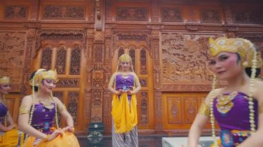 Mor bluzlar ve turuncu kuşaklarla geleneksel Bali kıyafetleri içinde beş dansçıdan oluşan bir grup, karmaşık şekilde oyulmuş ahşap paneller önünde poz verdiler. Dansçılar simetrik olarak düzenlenmiş zarif pozlarını ve ifadelerini sergiliyorlar..