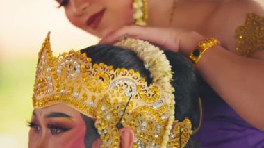 Geleneksel dans gösterisine hazırlanan iki kadının yakın çekimi. Bir kadın diğerinin süslü altın başlığını düzeltiyor. Makyaj ve mücevherleri var.. 