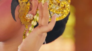 Geleneksel bir pozda, takılar ve renkli kıyafetlerle süslenmiş bir dansçının eli. Arka plan hafifçe bulanık, el hareketi ve hareketli kostümün karmaşık detaylarını vurguluyor..