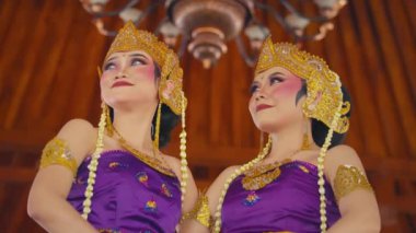 Geleneksel mor kostümler içinde altın aksesuarlar ve karmaşık mücevherlerle süslenmiş iki kadın dekoratif bir tavanın altında dans ediyorlar. Makyajları canlı, zarifçe poz verirken yüz ifadelerini vurguluyorlar..