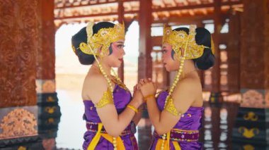 Geleneksel giyinmiş iki Balili dansçı. Karmaşık altın aksesuarlar ve canlı mor kostümlerle süslenmiş. Güzel dekore edilmiş ahşap bir çadırda yüz yüze duruyorlar..