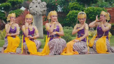 Beş kadından oluşan bir grup renkli kostümler içinde geleneksel bir dans gösterisi yapıyorlar. Altın aksesuarlar ve karmaşık saç şekilleriyle süslenmişler. Kültürel mirası sergiliyorlar. Arka planda yeşillik ve taş heykel yer alıyor..