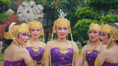 Geleneksel Bali kıyafetleri içinde mor elbiseler ve süslü altın aksesuarlardan oluşan beş kadından oluşan bir grup. Birlikte gür bir bahçe ortamında poz veriyorlar, karmaşık makyajlarını ve kültürel ifadelerini sergiliyorlar..