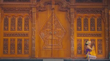 Karmaşık oymaları olan geleneksel bir ahşap kapı, canlı kostümlü bir dansçı için zemin görevi görüyor. Zarifçe oturan dansçı, kültürel giysiler sergiliyor, mimarinin ve dansın sanatını vurguluyor..