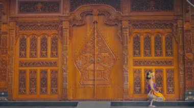 Geleneksel giyinmiş bir kadın karmaşık bir şekilde oyulmuş ahşap bir kapının önünde poz verir. Kapıda ayrıntılı desenler ve tasarımlar var. Kültürel sanatlar sergileniyor. Ortam canlı ve zengin bir mirası yansıtıyor..