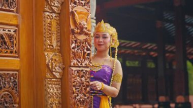 Geleneksel Bali kıyafeti giymiş bir kadın karmaşık bir şekilde oyulmuş ahşap bir kapının yanında duruyor. Altın bir taç ve mücevher takıyor, huzurlu bir ifadeyle, Bali kültürünün güzelliğini gösteriyor..