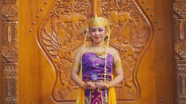 Geleneksel Bali kıyafetleri giymiş bir kadın. Üzerinde karmaşık altın aksesuarları olan mor ve sarı bir kıyafet var. Güzel oyulmuş ahşap bir arka fonun önünde duruyor, kültürel sanatını sergiliyor..