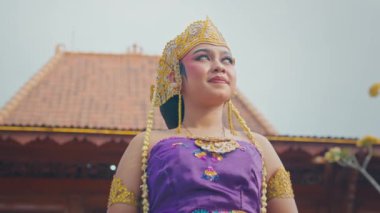 Geleneksel Bali kıyafeti giymiş, karmaşık mücevherler ve altın bir taç takmış genç bir kadın, açık havada güven içinde duruyor. Arka planda fayanslı çatısı olan geleneksel bir Bali binası var ve gökyüzü açık..