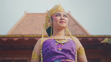 Geleneksel giysiler içinde, karmaşık mücevherler ve altın bir taç takmış mor bir elbise giyen genç bir kadın. Kendine güveni tam, sakin bir ifadeyle duruyor. Geleneksel mimarinin arka planına karşı..