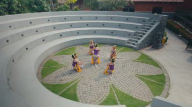 Renkli kostümlü beş kadının yer aldığı geleneksel bir dans gösterisi. Merkezi dansçı mor bir bluz ve sarı bir kuşak giyer, diğerleri ise benzer bir tavan arasında otururlar.