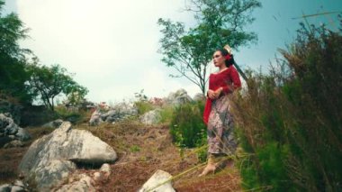 Geleneksel giyinmiş bir kadın yeşilliklerle çevrili kayalık bir arazide duruyor. Kırmızı bir bluz ve saçında çiçekler olan desenli bir etek giyiyor. Arka planda ağaçlar ve mavi bir gökyüzü var, sakin bir atmosfer yaratıyor..