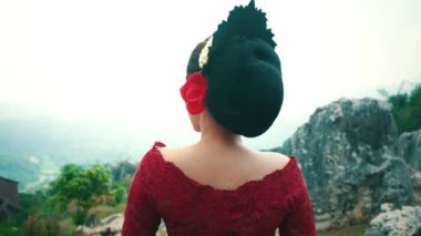 Kırmızı elbiseli bir kadın arkası kameraya dönük duruyor, manzaraya bakıyor. Onun saçı büyük siyah bir aksesuar ve kırmızı bir çiçekle zarif bir şekilde şekillendirilmiştir. Arka planda bulutlu bir gökyüzü altında kayalık arazi ve yeşillik yer alıyor..