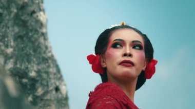 Renkli göz farı ve saçlarında kırmızı bir çiçek de dahil olmak üzere geleneksel makyajlı bir kadın. Manzaralı bir arkaplanda duruyor, özgüven ve zarafet saçıyor..