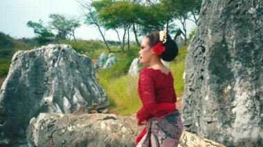 Geleneksel kıyafetli bir kadın doğal bir manzarada büyük bir kayanın üzerinde zarifçe poz verir. Kırmızı dantelli bir bluz ve yeşil ve kayalık oluşumlarla çevrili saçlarında bir çiçekle desenli bir sarong giyiyor..