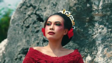 Kırmızı elbiseli bir kadın kayalık bir arka planda özgüvenle duruyor. Dramatik makyajı, canlı renkleri ve saçlarında çiçek aksesuarları var. Güçlü ve asil bir varlık sergiliyor..
