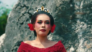 Çarpıcı makyajlı ve kırmızı elbiseli bir kadın kayalık bir arka planda duruyor. Dekoratif bir taç takıyor ve saçlarında çiçekler var. Canlı ve sanatsal bir tarzı var..