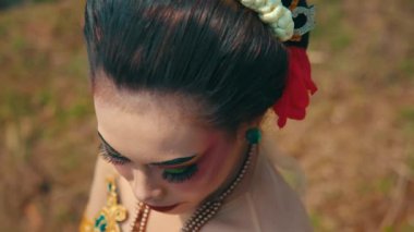 Dramatik makyajlı, canlı renklerde, kırmızı dudaklı ve geleneksel giyimli kalın göz makyajlı bir kişinin yakın plan portresi. Arkaplan bulanık, deneğin çarpıcı özelliklerini ve ifadesini vurguluyor.