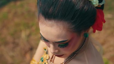 Dramatik makyajlı, kalın kaşları, renkli gözleri ve kırmızı dudakları olan bir kadının yakın plan portresi. Saçında geleneksel aksesuarlar ve çiçekler var, doğal bir arka plana göre dizilmiş..