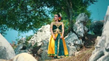 Kayalık arazinin yakınındaki çimenlerde çıplak ayakla yürüyen birinin yakın çekimi. Kişi renkli, desenli bir elbise giyiyor. Hareketle birlikte yavaşça akıyor.