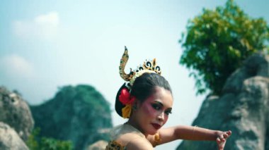 Geleneksel kıyafetli bir dansçı, doğal kaya oluşumları ve yeşilliklerle çevrili açık havada gösteri yapar. Dansçının arkası görünür rengarenk aksesuarlarla süslenmiştir. Saçında çiçekler vardır. Kültür mirası sergilenir..